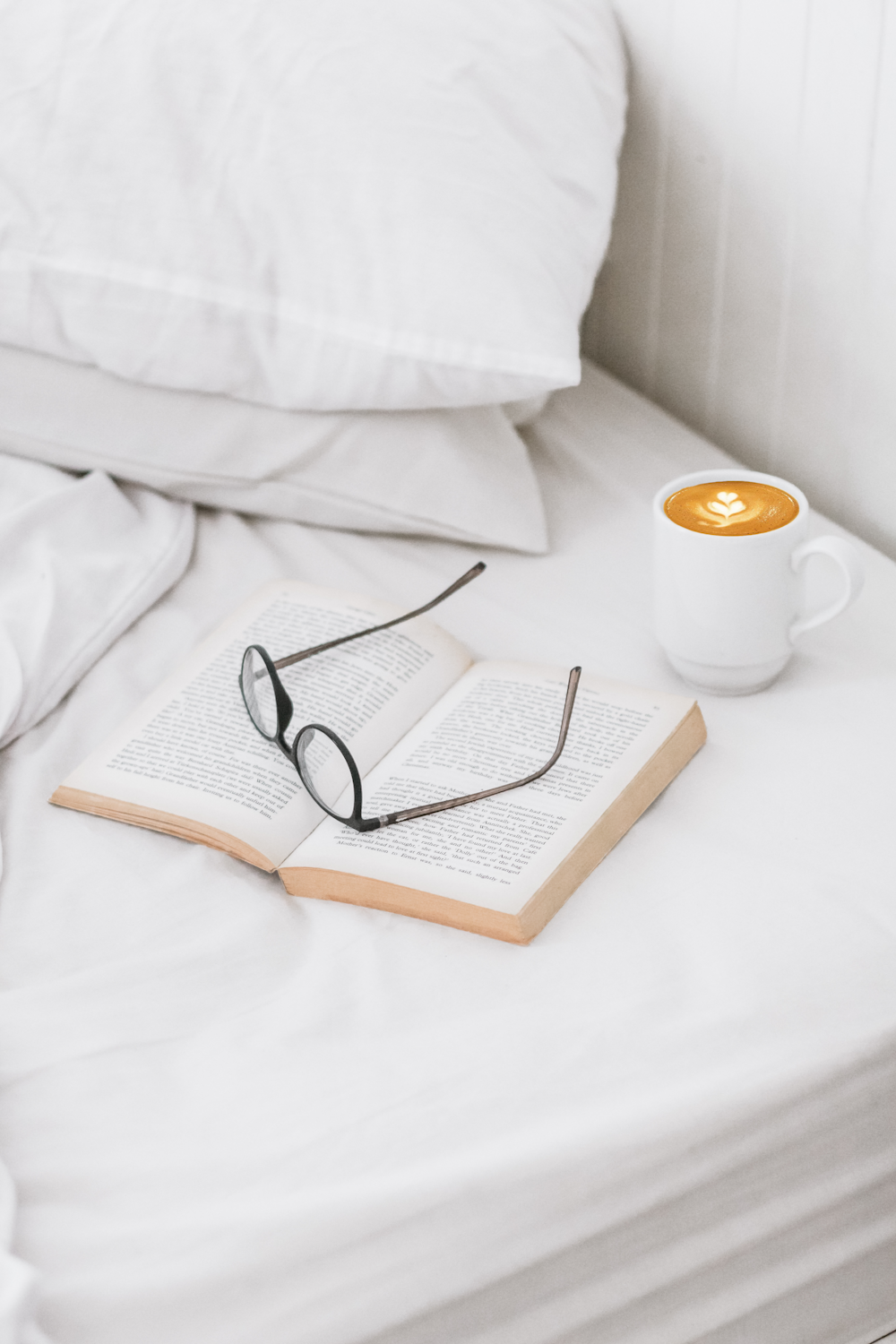 coffee on ceramic mug, book, and eyeglass on bed