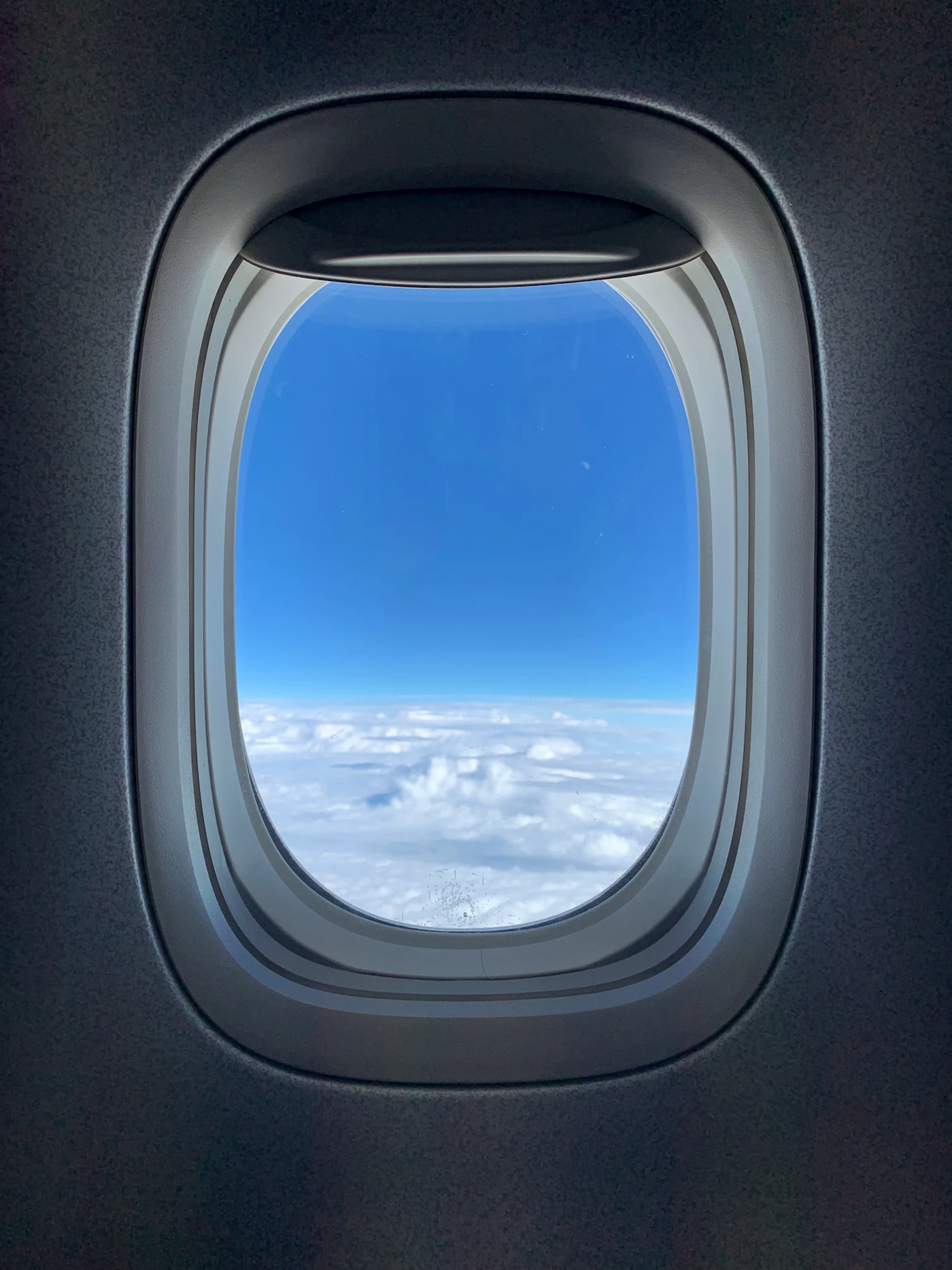 View through a cabin window of blue sky and a soft layer of clouds below during a smooth daytime flight