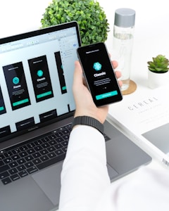 A person is holding a smartphone displaying app interface designs while sitting in front of a laptop with matching app designs on its screen. The setup is on a white desk that also has a plant, water bottle, and a book titled 'CEREAL'.