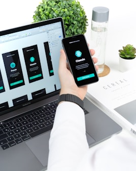 A person is holding a smartphone displaying app interface designs while sitting in front of a laptop with matching app designs on its screen. The setup is on a white desk that also has a plant, water bottle, and a book titled 'CEREAL'.