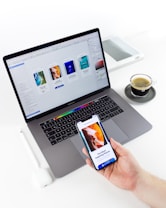 A person is holding a smartphone displaying a design app while sitting at a desk with a laptop, stylus, and cup of coffee. The laptop screen shows a design software interface with various templates and designs.