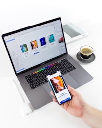 A person is holding a smartphone displaying a design app while sitting at a desk with a laptop, stylus, and cup of coffee. The laptop screen shows a design software interface with various templates and designs.