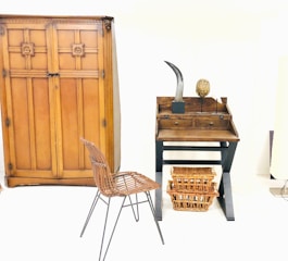A minimalist room featuring a large wooden wardrobe with decorative patterns. On the right, there's a small wooden desk with two drawers and items on top, including a sculpture resembling a horn and an oval-shaped object. Below the desk, two wicker baskets are placed. In front of the furniture, a metal frame chair with a woven seat adds to the rustic interior design.