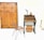 A minimalist room featuring a large wooden wardrobe with decorative patterns. On the right, there's a small wooden desk with two drawers and items on top, including a sculpture resembling a horn and an oval-shaped object. Below the desk, two wicker baskets are placed. In front of the furniture, a metal frame chair with a woven seat adds to the rustic interior design.