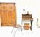 A minimalist room featuring a large wooden wardrobe with decorative patterns. On the right, there's a small wooden desk with two drawers and items on top, including a sculpture resembling a horn and an oval-shaped object. Below the desk, two wicker baskets are placed. In front of the furniture, a metal frame chair with a woven seat adds to the rustic interior design.