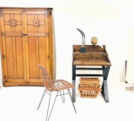 A minimalist room featuring a large wooden wardrobe with decorative patterns. On the right, there's a small wooden desk with two drawers and items on top, including a sculpture resembling a horn and an oval-shaped object. Below the desk, two wicker baskets are placed. In front of the furniture, a metal frame chair with a woven seat adds to the rustic interior design.