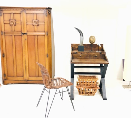 A minimalist room featuring a large wooden wardrobe with decorative patterns. On the right, there's a small wooden desk with two drawers and items on top, including a sculpture resembling a horn and an oval-shaped object. Below the desk, two wicker baskets are placed. In front of the furniture, a metal frame chair with a woven seat adds to the rustic interior design.