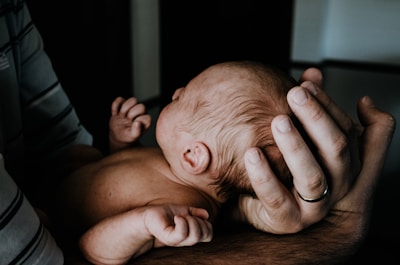 A newborn baby is gently cradled in an adult's arms, highlighting tenderness and protection. The adult's hand, wearing a ring, supports the baby's head. The baby's exposed back and slightly curled arms suggest a peaceful, resting position.
