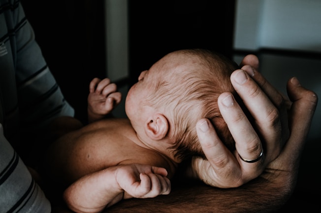 A newborn baby is gently cradled in an adult's arms, highlighting tenderness and protection. The adult's hand, wearing a ring, supports the baby's head. The baby's exposed back and slightly curled arms suggest a peaceful, resting position.