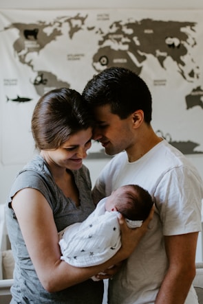 A tender moment between a couple holding their newborn baby. The parents are standing close together with their foreheads touching, gazing lovingly at the sleeping infant wrapped in a white blanket with star patterns. In the background, a world map is partially visible.