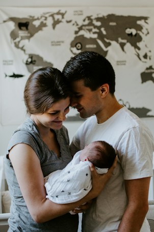 A tender moment between a couple holding their newborn baby. The parents are standing close together with their foreheads touching, gazing lovingly at the sleeping infant wrapped in a white blanket with star patterns. In the background, a world map is partially visible.