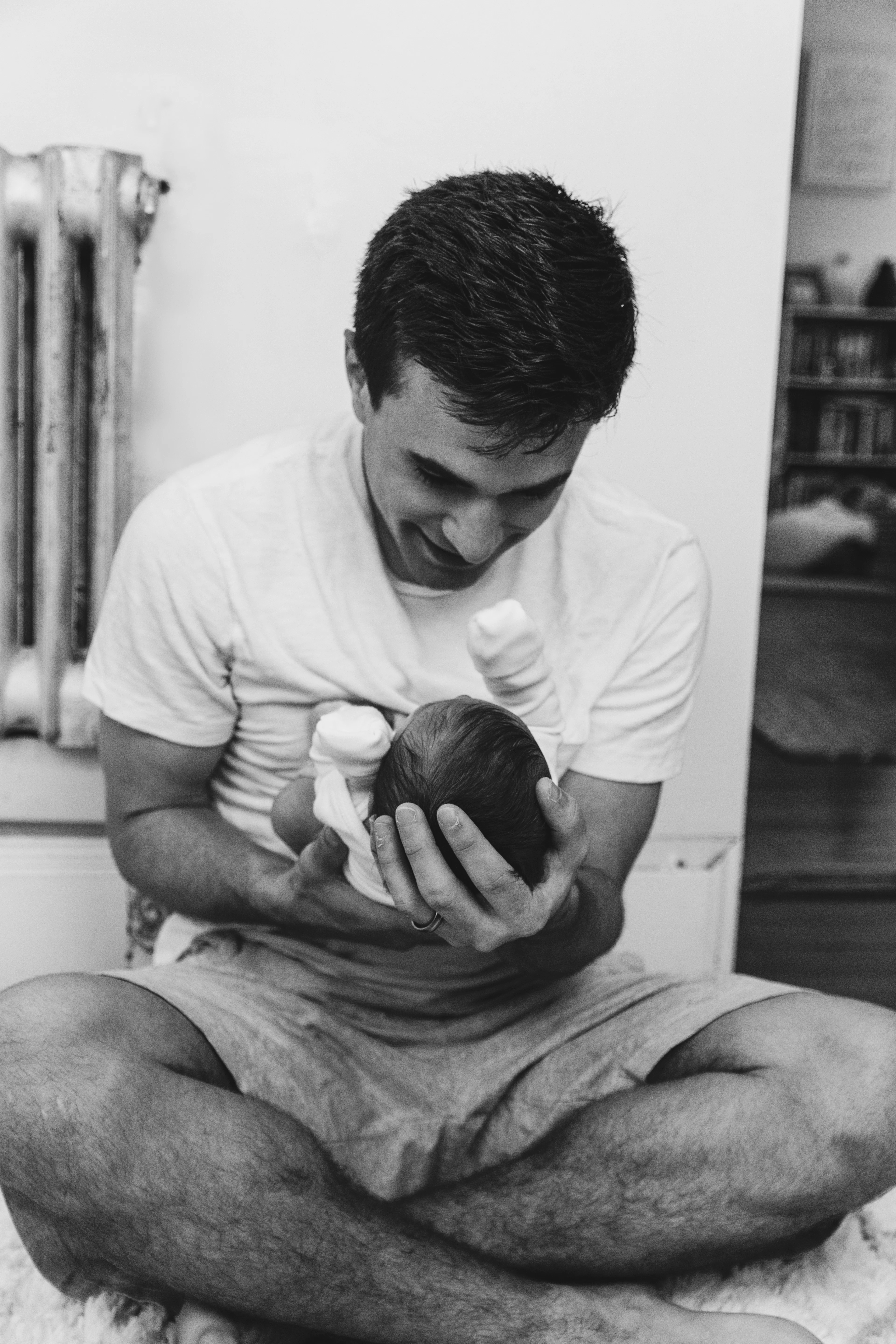 Man sitting on a bed cradling a newborn, in black and white
