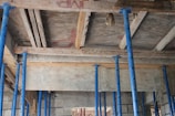 A construction site with several blue metal poles supporting a wooden ceiling. The ceiling consists of wooden planks and beams with a rough, unfinished appearance. Some bricks and concrete blocks are visible in the background.