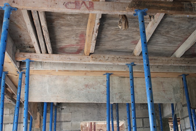 A construction site with several blue metal poles supporting a wooden ceiling. The ceiling consists of wooden planks and beams with a rough, unfinished appearance. Some bricks and concrete blocks are visible in the background.