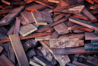 A pile of assorted wooden planks and pieces stacked unevenly, with various textures and colors. Some wood appears weathered with visible grain patterns, while others are smooth and freshly cut. The planks are in different shades of brown, some with hints of green and orange.
