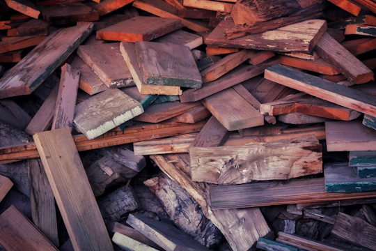 A pile of assorted wooden planks and pieces stacked unevenly, with various textures and colors. Some wood appears weathered with visible grain patterns, while others are smooth and freshly cut. The planks are in different shades of brown, some with hints of green and orange.