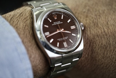A close-up view of a luxury wristwatch with a metallic silver band and a dark brown dial, worn on a wrist. The watch face displays the brand name and other details clearly.