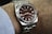 A close-up view of a luxury wristwatch with a metallic silver band and a dark brown dial, worn on a wrist. The watch face displays the brand name and other details clearly.