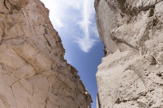 A narrow gap between two massive rock formations under a bright blue sky, with light, wispy clouds overhead. The rocks show a variety of textures and shades of beige, with weathered, uneven surfaces.