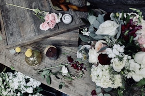 A rustic wooden table adorned with a variety of fresh flowers including roses and hydrangeas in soft pink, white, and burgundy tones. A small bouquet of pink roses lies on one corner, and a vase with yellow spherical flowers is placed nearby. Wooden boards and a small candle create a textured, natural backdrop.