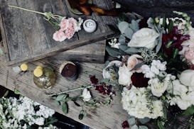 A rustic wooden table adorned with a variety of fresh flowers including roses and hydrangeas in soft pink, white, and burgundy tones. A small bouquet of pink roses lies on one corner, and a vase with yellow spherical flowers is placed nearby. Wooden boards and a small candle create a textured, natural backdrop.