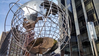 A large metallic globe sculpture with a network of lines representing longitude and latitude stands prominently against a backdrop of modern, reflective glass buildings. Sunlight casts bright reflections off the metal, emphasizing the intricate design of the sculpture. Tall urban structures surround the globe, capturing a sense of city life and architectural diversity.