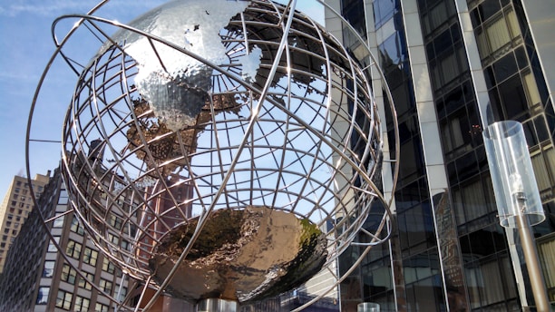 A large metallic globe sculpture with a network of lines representing longitude and latitude stands prominently against a backdrop of modern, reflective glass buildings. Sunlight casts bright reflections off the metal, emphasizing the intricate design of the sculpture. Tall urban structures surround the globe, capturing a sense of city life and architectural diversity.