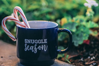 A navy blue mug with the phrase 'Snuggle Season' printed in white sits on a surface. Two candy canes are placed inside the mug, adding a festive touch. The background is softly out of focus, featuring green foliage and a hint of white flowers.