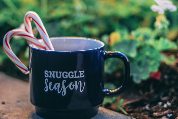 A navy blue mug with the phrase 'Snuggle Season' printed in white sits on a surface. Two candy canes are placed inside the mug, adding a festive touch. The background is softly out of focus, featuring green foliage and a hint of white flowers.
