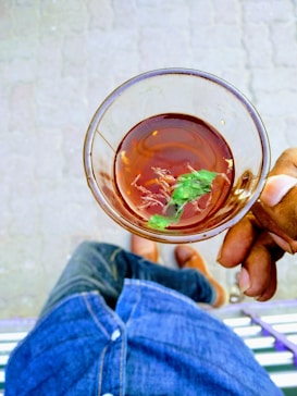 A person is holding a glass of tea with herbs in it, viewed from above. The person's hand is visible holding the cup, with denim blue jeans and brown shoes in the background. The ground has a concrete pavement pattern.