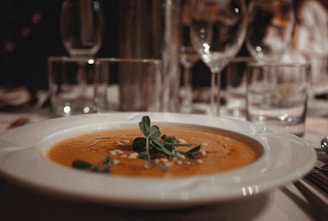 A close-up of a bowl of creamy soup garnished with green herbs and seeds, placed on a table with elegantly set glassware and utensils in the background.