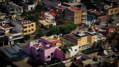 A densely packed urban area featuring a variety of residential buildings with flat rooftops. The architecture includes colorful facades with shades of pink, yellow, and earth tones. Solar panels are visible on some roofs, and there are trees and power lines interspersed between the buildings.