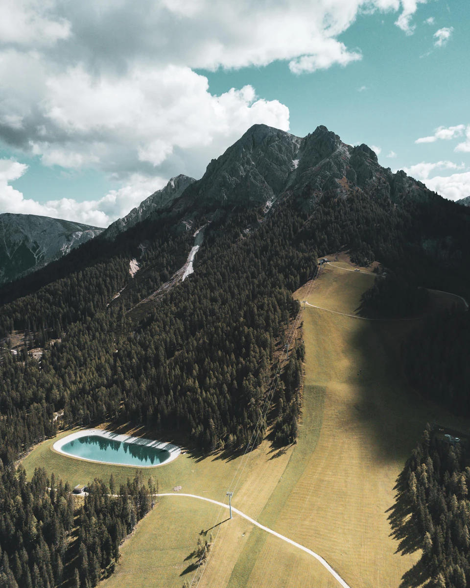 The image presents an aerial view of a mountainous landscape, with a large mountain range stretching across the horizon and its peak shrouded in clouds. The mountains are densely covered in trees, creating a lush green canopy that contrasts with the gray sky above. Nestled amidst this verdant expanse is a small lake, its surface reflecting the surrounding greenery. The lake is encircled by a winding path, inviting exploration of the natural beauty within.