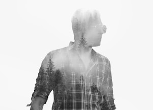 A monochromatic artistic composition blends a man wearing sunglasses and a plaid shirt with silhouettes of tall trees overlapping his form. The image creates a double exposure effect, merging human and nature elements seamlessly.