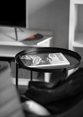 A minimalist interior setting with a black circular tray table. On the table lies an open magazine with the title 'Bauhaus No. 9,' featuring a monochrome image. The background shows a blurry TV off to one side on a white cabinet, and the corner of a black chair is visible in the foreground.