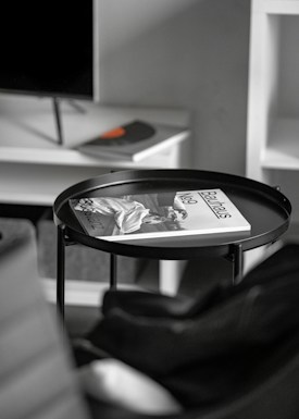 A minimalist interior setting with a black circular tray table. On the table lies an open magazine with the title 'Bauhaus No. 9,' featuring a monochrome image. The background shows a blurry TV off to one side on a white cabinet, and the corner of a black chair is visible in the foreground.