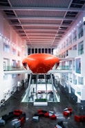 A modern indoor space featuring a bright red, futuristic pod structure elevated on stilts at the center, surrounded by glass balconies and white walls. Geometric furniture pieces in red, black, and gray are scattered on a polished dark floor, contributing to a minimalistic and contemporary aesthetic.