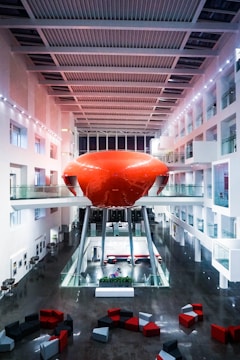 A modern indoor space featuring a bright red, futuristic pod structure elevated on stilts at the center, surrounded by glass balconies and white walls. Geometric furniture pieces in red, black, and gray are scattered on a polished dark floor, contributing to a minimalistic and contemporary aesthetic.