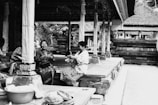 Two women are sitting on a platform in a traditional setting, engaged in conversation and sharing a moment of laughter. They are dressed in cultural attire, surrounded by intricately decorated columns and wooden architecture.