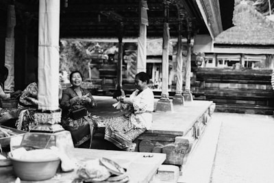 Two women are sitting on a platform in a traditional setting, engaged in conversation and sharing a moment of laughter. They are dressed in cultural attire, surrounded by intricately decorated columns and wooden architecture.