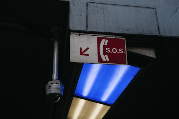 An S.O.S. sign with a red background and white telephone symbol is mounted on a wall. Near the sign, a surveillance camera is positioned, and a blue fluorescent light is visible below.