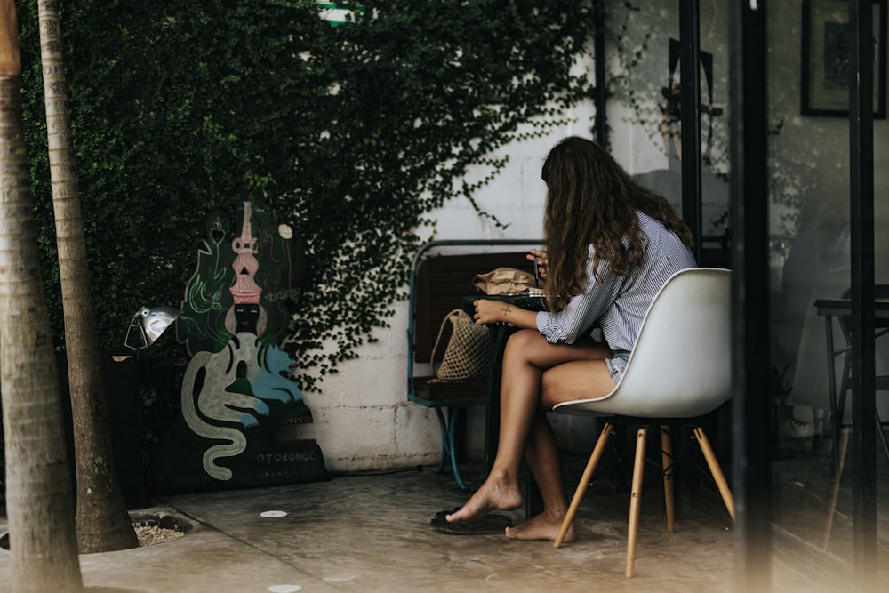 Mujer vestida con camisa gris de manga 3/4 sentada en una silla blanca frente a una mesa negra cerca del árbol