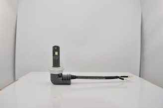 A small electronic device with a rectangular black body and a white circular base is connected to a black cable. The setup is placed on a smooth, white surface against a light gray background.