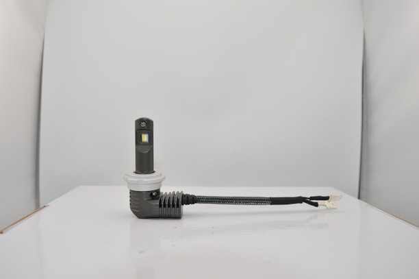 A small electronic device with a rectangular black body and a white circular base is connected to a black cable. The setup is placed on a smooth, white surface against a light gray background.