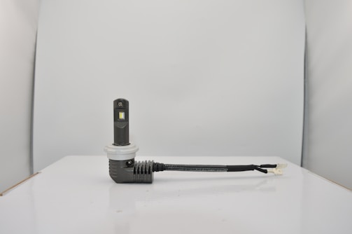 A small electronic device with a rectangular black body and a white circular base is connected to a black cable. The setup is placed on a smooth, white surface against a light gray background.