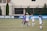 A group of female soccer players are actively engaged in a sports match on a grass field. Two players, one in a white uniform and the other in a blue uniform, are in the foreground leaping to head the ball. Several other players are positioned nearby, all appearing focused and in motion. The background features a blue banner with text, and a building with large windows and a black fence.