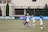 A group of female soccer players are actively engaged in a sports match on a grass field. Two players, one in a white uniform and the other in a blue uniform, are in the foreground leaping to head the ball. Several other players are positioned nearby, all appearing focused and in motion. The background features a blue banner with text, and a building with large windows and a black fence.