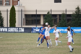 A group of female soccer players are actively engaged in a sports match on a grass field. Two players, one in a white uniform and the other in a blue uniform, are in the foreground leaping to head the ball. Several other players are positioned nearby, all appearing focused and in motion. The background features a blue banner with text, and a building with large windows and a black fence.