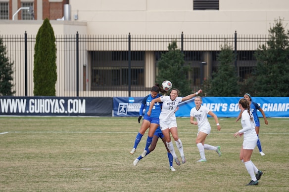 A group of female soccer players are actively engaged in a sports match on a grass field. Two players, one in a white uniform and the other in a blue uniform, are in the foreground leaping to head the ball. Several other players are positioned nearby, all appearing focused and in motion. The background features a blue banner with text, and a building with large windows and a black fence.