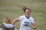A female soccer player wearing a white jersey with 'Hoyas' and the number 23 printed on it is captured mid-motion, smiling with hair flying back. She appears to be on a grass field, engaging with someone in the foreground who wears a jacket with a fur collar.